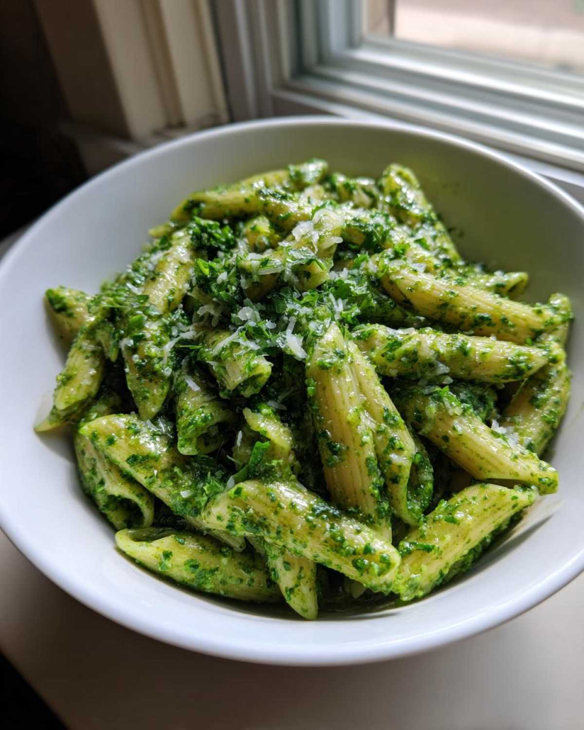 A close-up shot of penne pasta coated in vibrant green creamy pesto pasta, topped with grated Parmesan cheese.