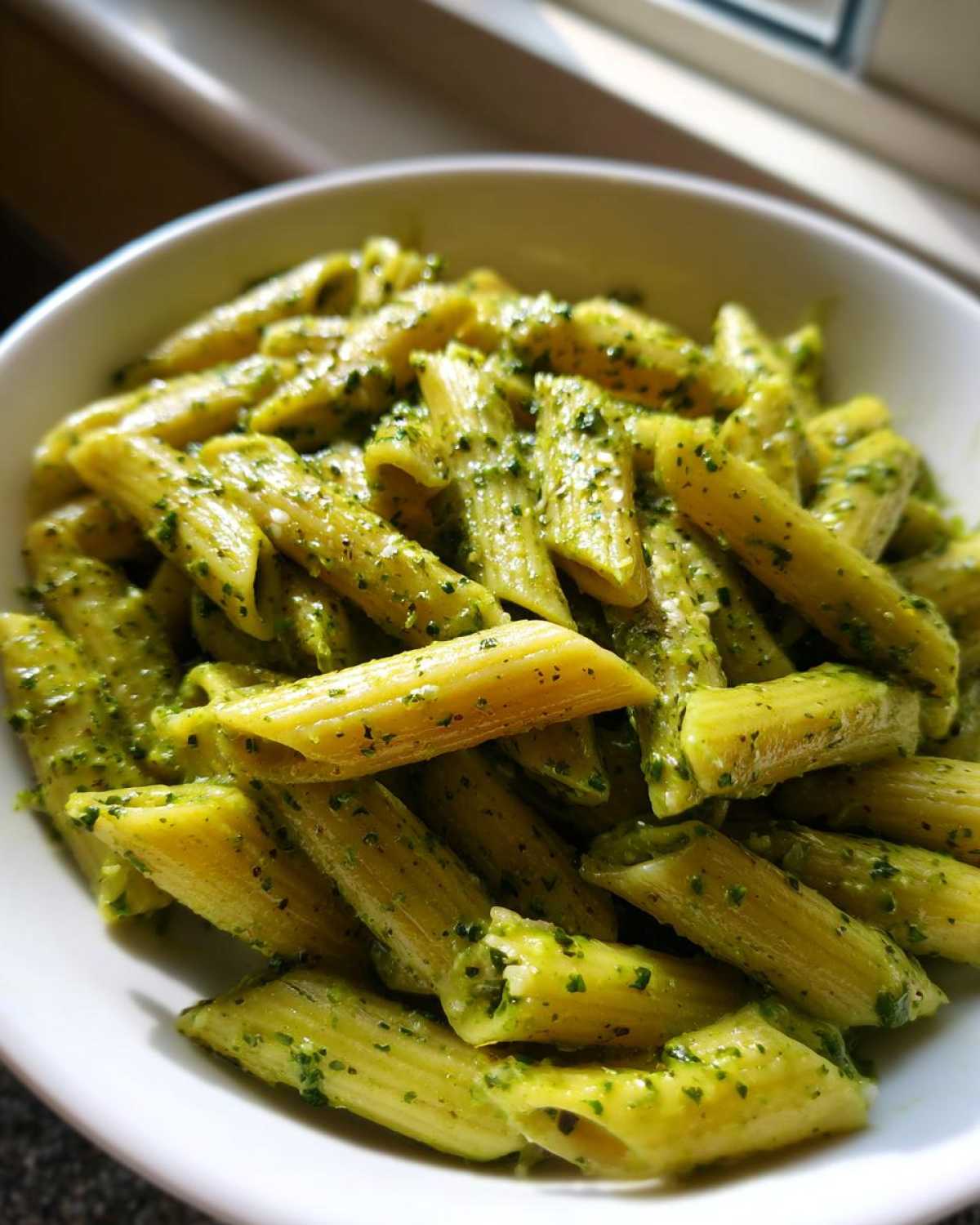 Close-up of penne pasta coated generously in bright green creamy pesto pasta sauce in a white bowl.