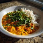 A vibrant bowl of creamy orange pumpkin curry served alongside white rice, topped generously with fresh cilantro and chili flakes.