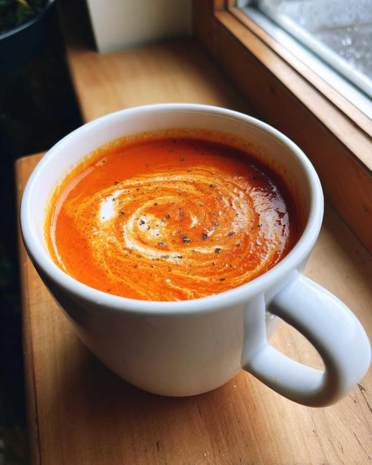 Close-up of a rich, orange tomato basil soup in a white mug, swirled with cream and sprinkled with pepper.
