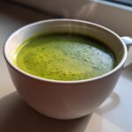 Close-up of a white mug filled with hot, vibrant green zucchini soup, showing steam rising.