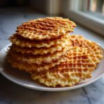 A stack of freshly made, golden brown, crisp pizzelle cookies resting on a white plate.