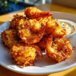 A pile of golden brown, crispy coconut shrimp recipe pieces served on a white plate with a small dipping sauce bowl.