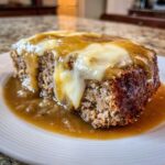 A close-up of a slice of crockpot french onion meatloaf topped with melted white cheese and rich brown gravy.