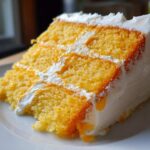 A close-up of a moist, three-layer slice of Creamsicle Cake with bright orange sponge and white frosting.