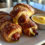 Three freshly baked pretzel dogs sprinkled with coarse salt, served with a side of bright yellow cheese sauce.