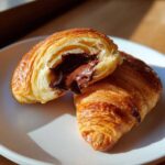 Close-up of a flaky chocolate croissant cut in half, showing gooey melted chocolate filling.
