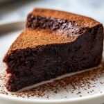 A close-up of a rich, dark slice of flourless chocolate torte dusted generously with cocoa powder.