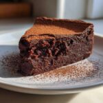 A decadent slice of flourless chocolate torte, intensely dark and fudgy, dusted with cocoa powder on a white plate.