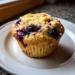 Close-up of a single, fluffy blueberry cottage cheese muffin topped with golden streusel, resting on a white plate.