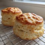 Two tall, fluffy scones with golden-brown tops cooling on a wire rack near a window.