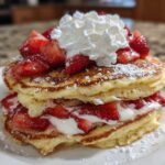 A tall stack of fluffy strawberry pancakes layered with cream and topped with fresh strawberries and whipped cream.