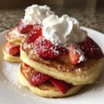 Close-up of fluffy strawberry pancakes stacked high, topped with fresh strawberries, whipped cream, and powdered sugar.