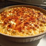 A freshly baked, creamy elote dip topped with browned cheese and paprika in a light tan baking dish.