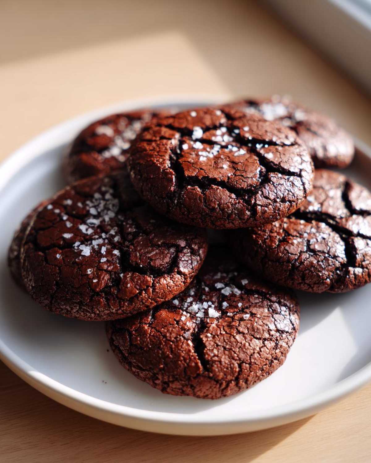 A stack of rich, dark chocolate brownie cookies topped with flaky sea salt on a white plate.