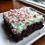 A close-up of a fudgy peppermint brownie topped with light green mint frosting and crushed candy canes.