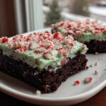 Two slices of fudgy peppermint brownies topped with light green mint frosting and crushed candy canes.