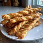 A close-up of a pile of golden, flaky, twisted cheese straws recipe on a white plate.