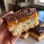 A person holding a cross-section of a delicious twix cookies bar showing the shortbread base, thick caramel layer, and chocolate topping.