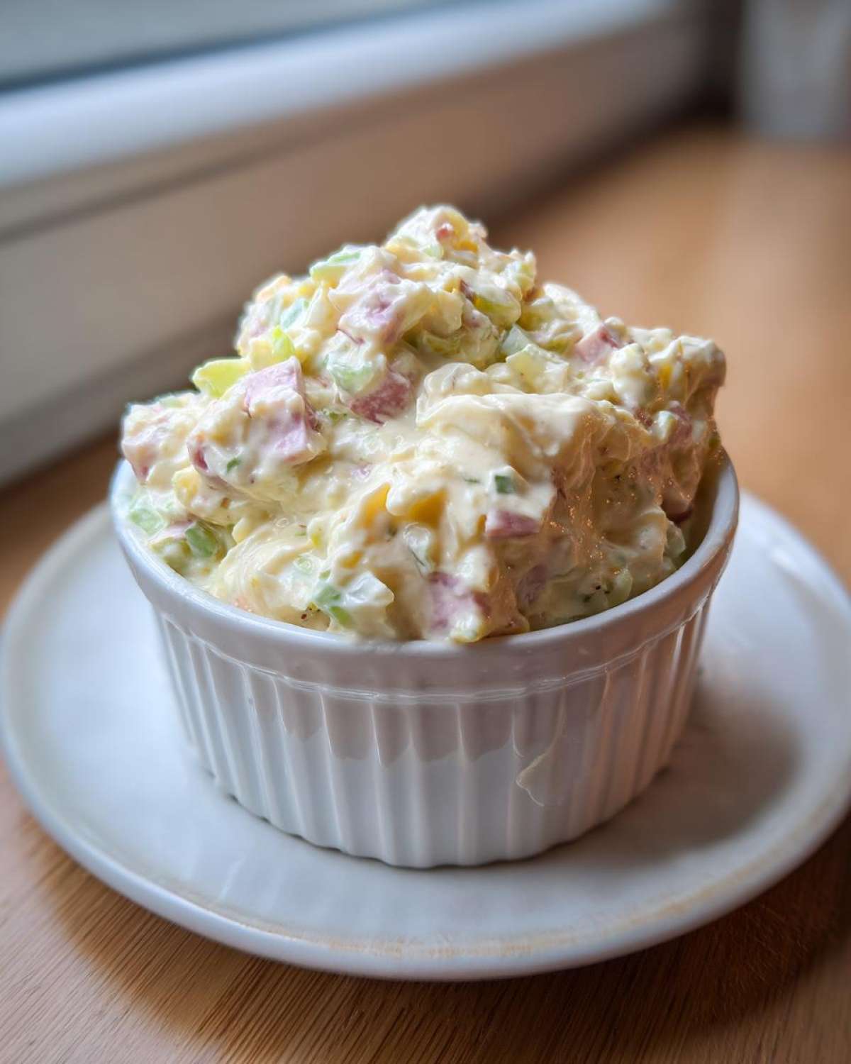 A close-up of a creamy portion of ham salad recipe served in a white fluted ramekin on a small plate.