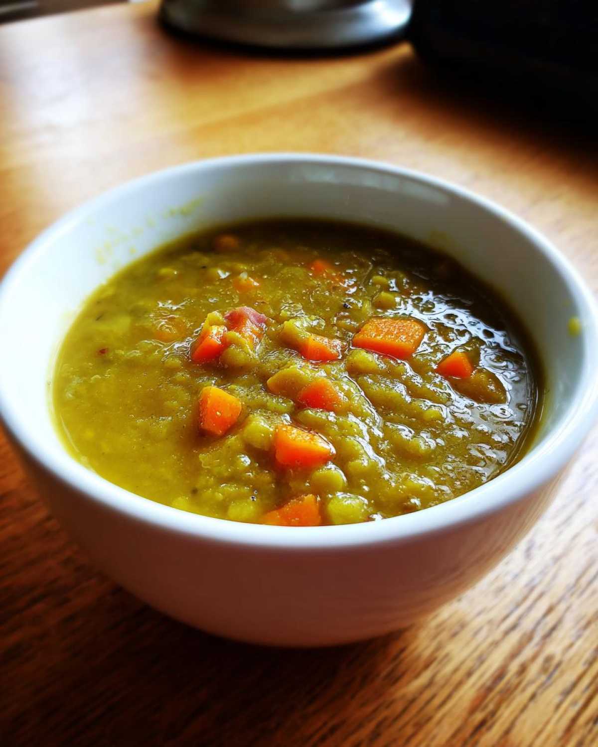 Close-up of a thick, green pea soup topped with visible chunks of bright orange carrots in a white bowl.
