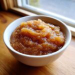 A close-up of chunky, homemade applesauce served in a white bowl on a wooden surface.