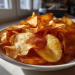 Close-up of a white bowl overflowing with golden, crispy homemade potato chips seasoned with paprika.