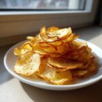 A tall pile of golden, homemade potato chips seasoned with herbs, served on a white plate.