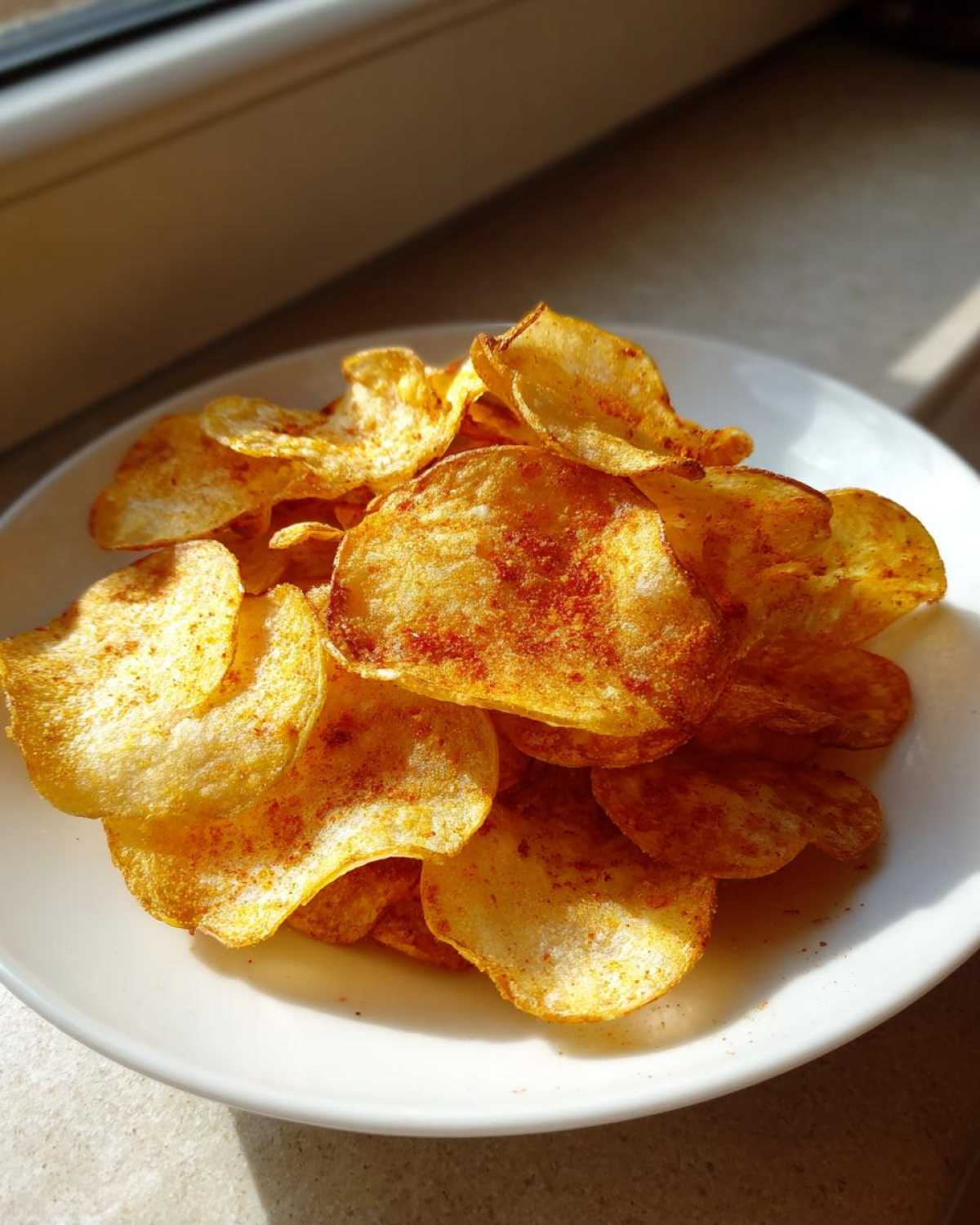 A pile of golden, crispy homemade potato chips seasoned with paprika, served on a white plate.