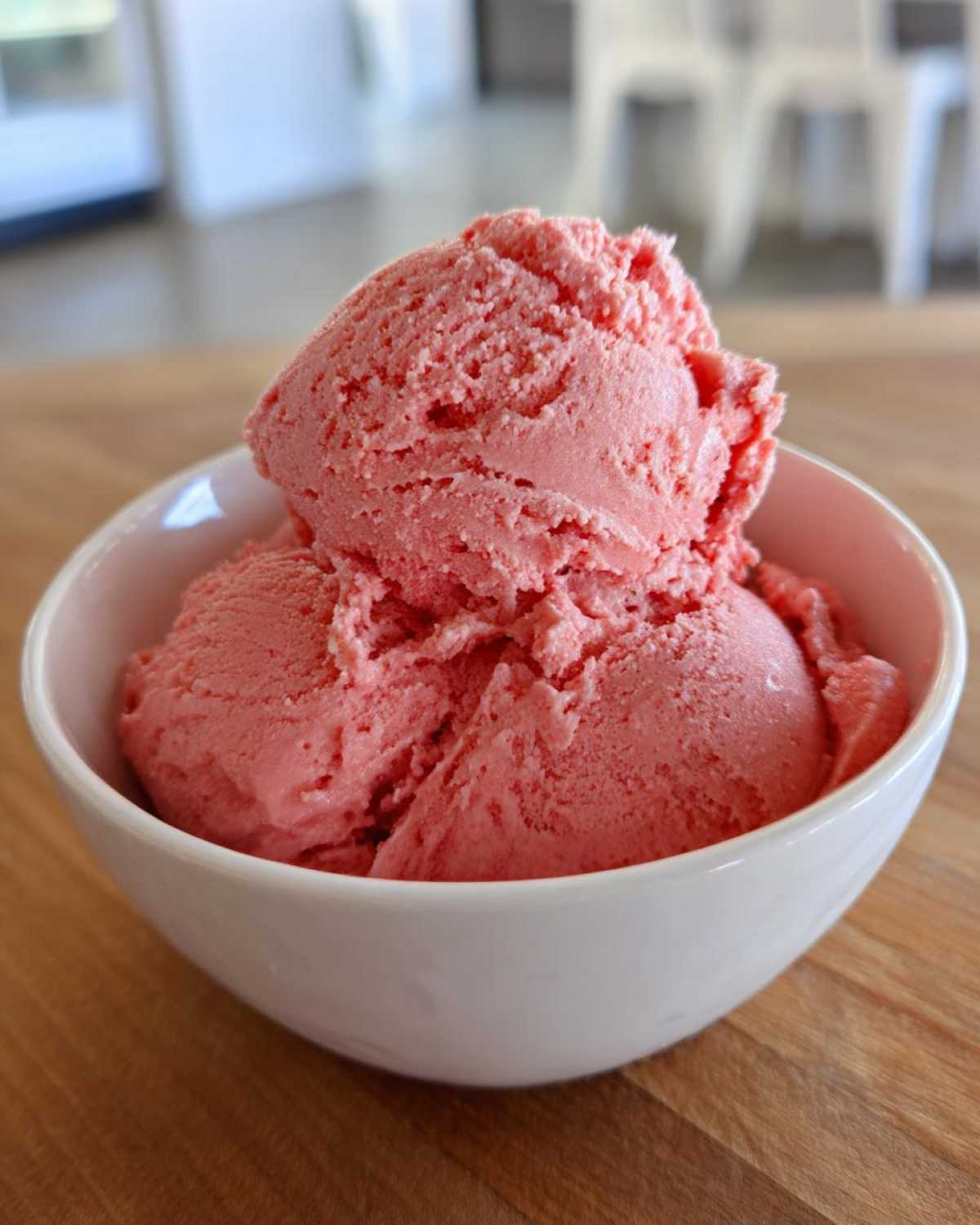 Three generous scoops of bright pink strawberry ice cream served in a white bowl on a wooden surface.