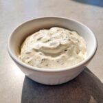 A close-up of creamy, homemade tartar sauce flecked with green herbs, served in a small white bowl.