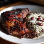 Close-up of a juicy, charred piece of Jamaican jerk chicken served next to rice and red kidney beans.