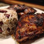 Close-up of two pieces of charred, glazed Jamaican Jerk Chicken served next to a mound of rice and peas.
