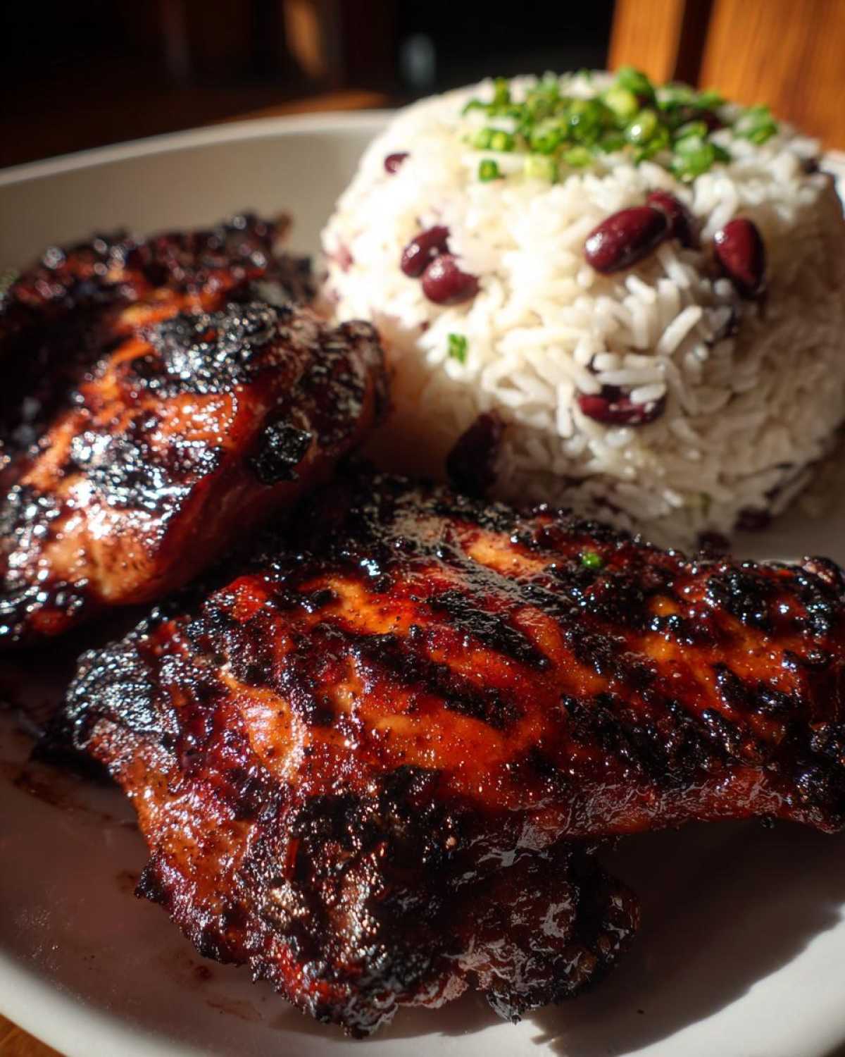 Two pieces of dark, glazed Jamaican jerk chicken served next to a mound of rice and peas topped with green onions.