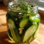 Close-up of sliced cucumbers, dill, garlic, and peppercorns in a jar of brine for refrigerator pickles.
