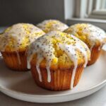 Four bright yellow lemon poppy seed muffins topped with thick white glaze and poppy seeds, resting on a light plate.