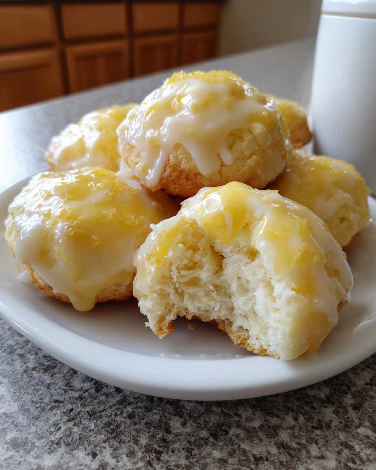A stack of soft, glazed lemon ricotta cookies on a white plate, one cookie has a bite taken out showing the fluffy interior.