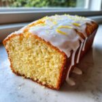 Close-up of a moist lemon loaf cake, sliced to show texture, topped with white glaze and lemon zest.