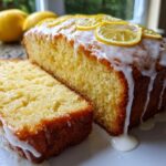 A moist lemon loaf cake, partially sliced, covered in white glaze and topped with candied lemon slices.