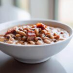 Close-up of a white bowl filled with creamy black eyed peas topped with chunks of smoked ham.