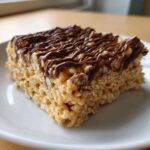 A close-up of a square peanut butter rice krispie treats drizzled generously with melted chocolate.