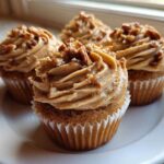 Four delicious pecan pie cupcakes with swirled brown sugar frosting and chopped pecans on a white plate.