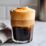 A tall glass of rich pumpkin cream cold brew topped with thick, frothy pumpkin foam and a sprinkle of spice.