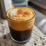 Close-up of a glass filled with pumpkin cream cold brew, showing dark coffee below and thick orange foam topped with cinnamon.