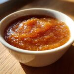 Close-up of rich, brown homemade applesauce served in a small white bowl, highlighted by sunlight.