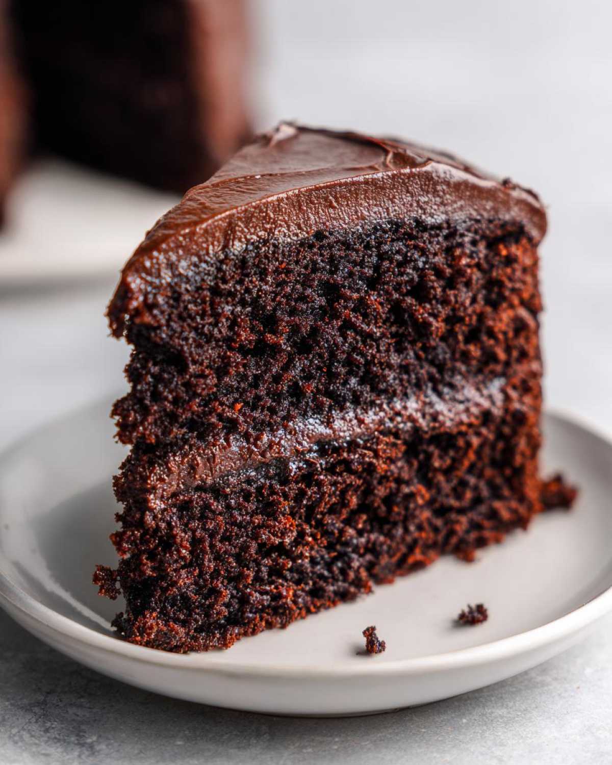 A moist, dark slice of chocolate cake recipe with thick, glossy chocolate frosting on a light gray plate.