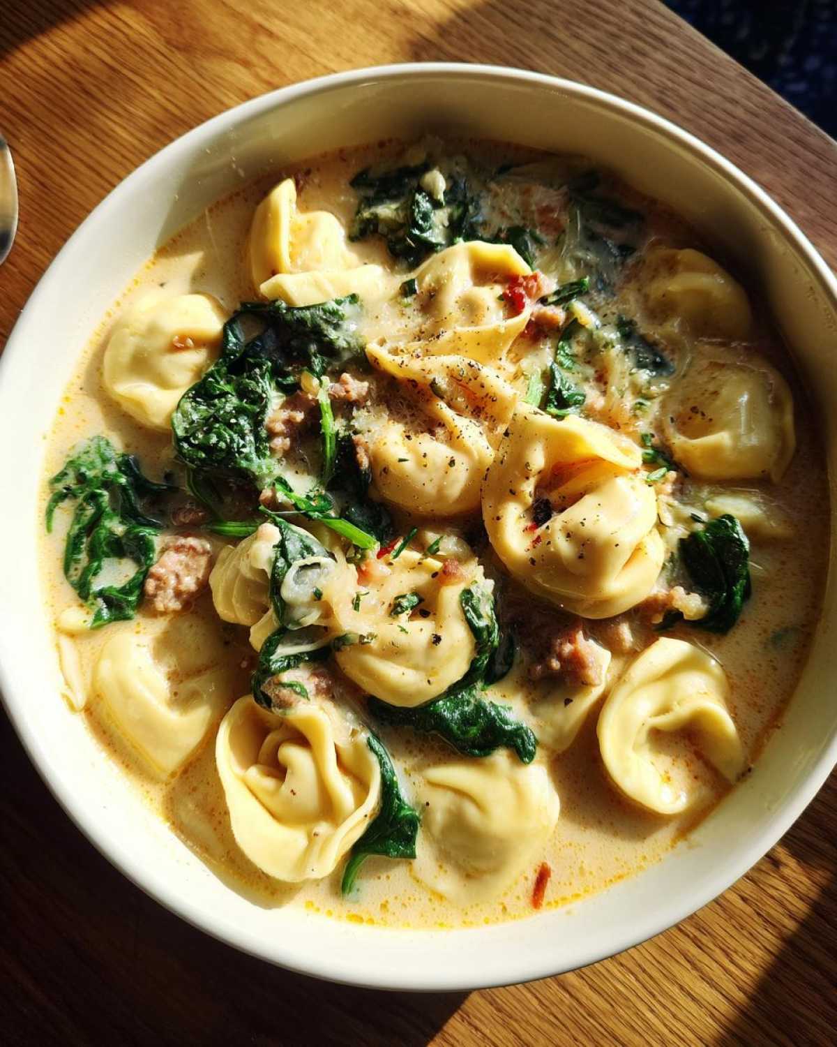Top-down view of a bowl of creamy sausage tortellini soup with wilted spinach, seasoned with pepper.