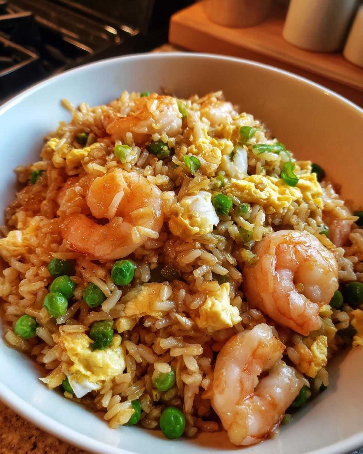 Close-up of a white bowl filled with flavorful shrimp fried rice, featuring large pink shrimp, scrambled egg, and bright green peas.