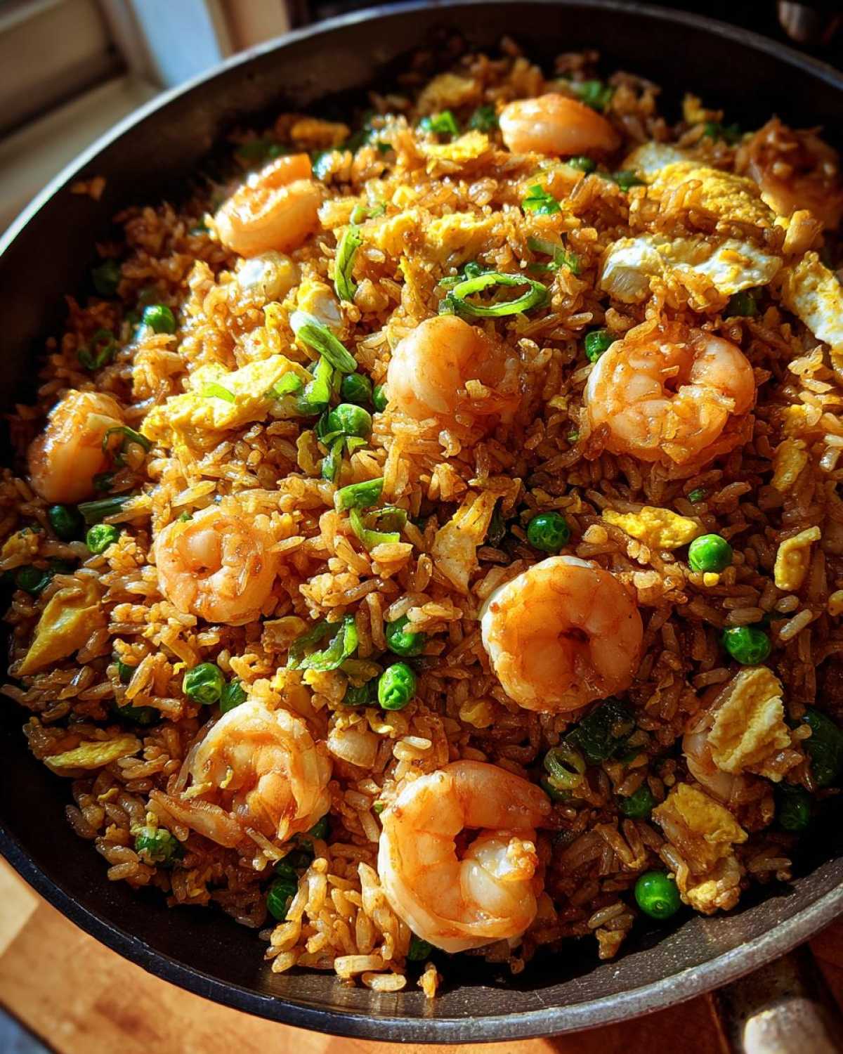 A close-up overhead view of freshly made shrimp fried rice with large shrimp, peas, egg, and green onions in a dark skillet.