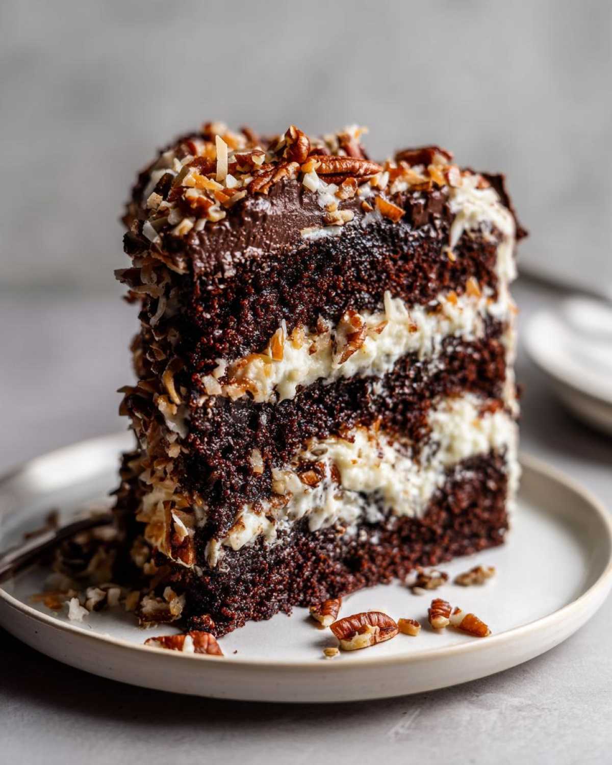 A tall, moist slice of german chocolate cake featuring dark layers, white coconut-pecan frosting, and topped with toasted pecans.